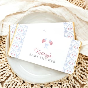 May include: A rectangular candy bar wrapper with a white background, featuring a repeating blue lattice pattern with small bows on the sides. The center displays two small American flags tied with a blue bow, and the text Kelsey's BABY SHOWER.