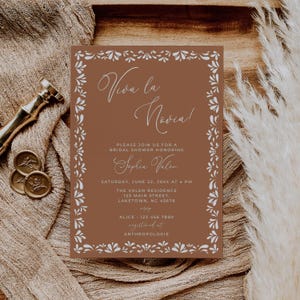 Puede incluir: Una tarjeta de invitación marrón con borde floral blanco y texto. El texto dice "Viva la Novia!" y detalla una despedida de soltera en honor a "Sephia Valen". Incluye fecha, hora y lugar. También se proporciona información de RSVP.