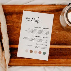 May include: A wedding details card with the text "the details" in elegant script. The card includes information about accommodation, transportation, and dress code, with a color palette at the bottom. The card is on a wooden surface.