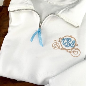 May include: A white quarter-zip sweatshirt featuring a silver metal zipper pull with a light blue polka dot ribbon attached. The chest displays an embroidered tan pumpkin carriage design with blue monogrammed letters inside the carriage frame.
