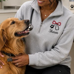 Puede incluir: Sudadera gris claro con cremallera de cuarto de cierre con un diseño de estetoscopio negro, huella de pata y lazo rojo. El nombre "Sharon" está bordado debajo. Un perro golden retriever está en primer plano.
