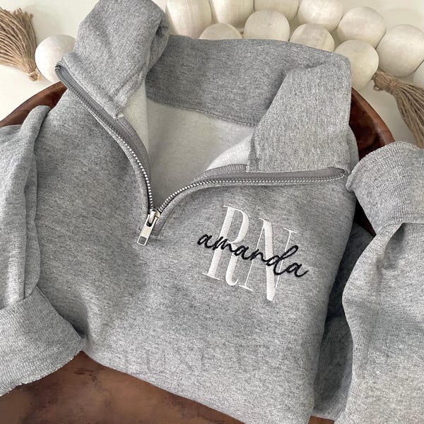 May include: A heather gray quarter-zip sweatshirt featuring a silver metal zipper and a high collar. The chest displays white embroidered block letters RN overlaid with the name amanda in black cursive script embroidery.
