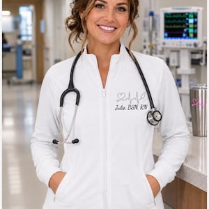 May include: A white long-sleeve zip-up jacket featuring an embroidered design on the left chest with a heart, an EKG heartbeat line, a stethoscope icon, and the text "Julie, BSN, RN" in black thread. A black stethoscope rests around the neck.