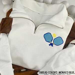 May include: White pullover sweatshirt with a quarter-zip neckline. The sweatshirt features an embroidered design of two blue pickleball paddles and a yellow ball. The text at the bottom of the image reads: THREAD COLORS: MONACO BLUE, CIEL, BUFF AND YELLOW.