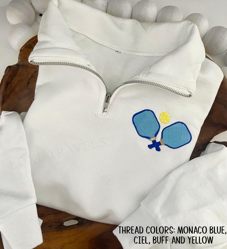 May include: White pullover sweatshirt with a quarter-zip neckline. The sweatshirt features an embroidered design of two blue pickleball paddles and a yellow ball. The text at the bottom of the image reads: THREAD COLORS: MONACO BLUE, CIEL, BUFF AND YELLOW.