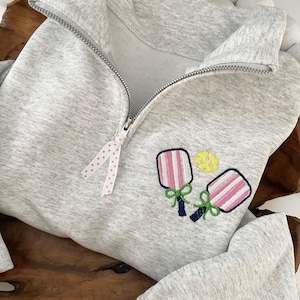 May include: A light gray zip-up sweatshirt with a white and pink polka dot ribbon on the zipper. The sweatshirt features embroidered pickleball paddles and a yellow ball.