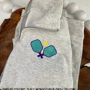 May include: A light gray quarter-zip sweatshirt with an embroidered pickleball design. The design features two teal paddles and a yellow ball, with red and blue accents. The text at the bottom reads "THREAD COLORS: TEAL/ MONACO BLUE/RED/ YELLOW".