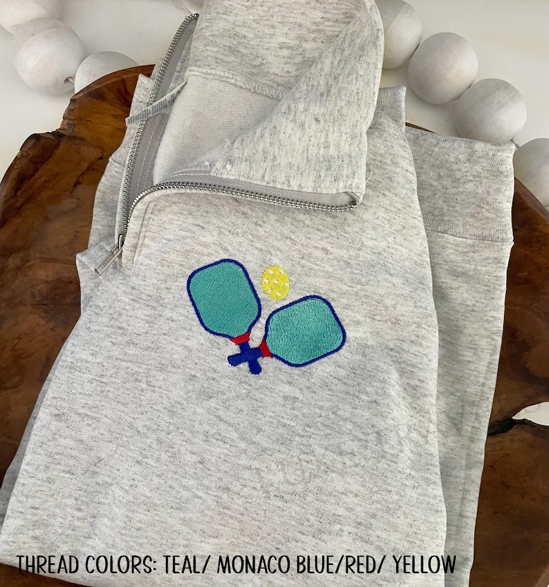 May include: A light gray quarter-zip sweatshirt with an embroidered pickleball design. The design features two teal paddles and a yellow ball, with red and blue accents. The text at the bottom reads "THREAD COLORS: TEAL/ MONACO BLUE/RED/ YELLOW".