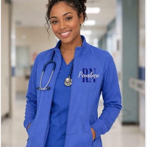 May include: A royal blue zip-up jacket featuring a mock neck, long sleeves, and two front pockets. The left chest is embroidered with the name Penelope in white script, layered over the dark blue letters RN in a bold, block font style.