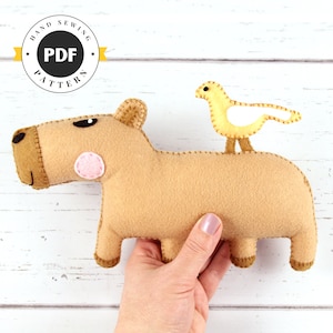May include: A tan felt capybara plush toy with a yellow bird perched on its back. The capybara has a pink cheek and brown stitching. The image includes a circular logo with the text "HAND SEWING PATTERN PDF".
