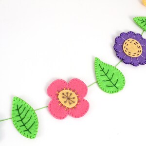 May include: A colorful felt flower garland with pink, purple, and yellow flowers, and green leaves. The flowers and leaves are connected by a green string. The garland is a decorative item.