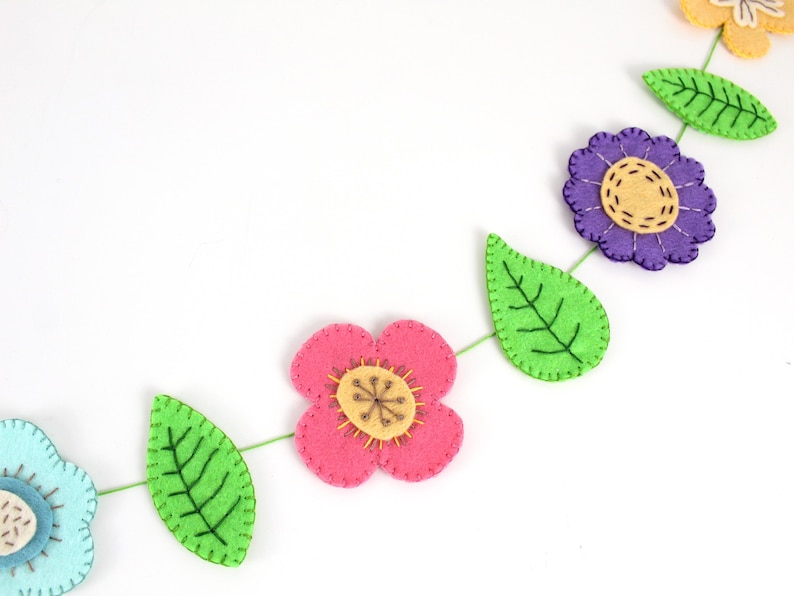 May include: A colorful felt flower garland with pink, purple, and yellow flowers, and green leaves. The flowers and leaves are connected by a green string. The garland is a decorative item.