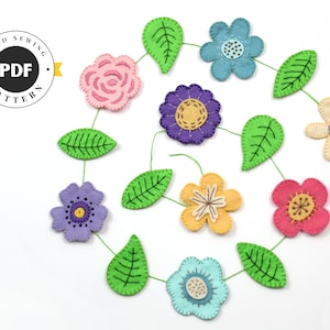 Op de afbeelding: Een kleurrijke vilten bloemenslinger met diverse bloemdesigns in roze, blauw, paars en geel, verbonden door groene stelen en bladeren. De afbeelding bevat een cirkelvormig logo met de tekst "HAND SEWING PDF PATTERN".