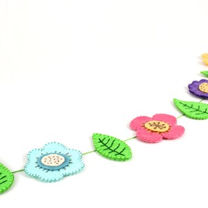 May include: A felt flower garland featuring a variety of colorful flowers and green leaves. The flowers are in shades of pink, blue, purple, and yellow, with detailed stitching and wooden center accents. The garland is strung on a green thread.