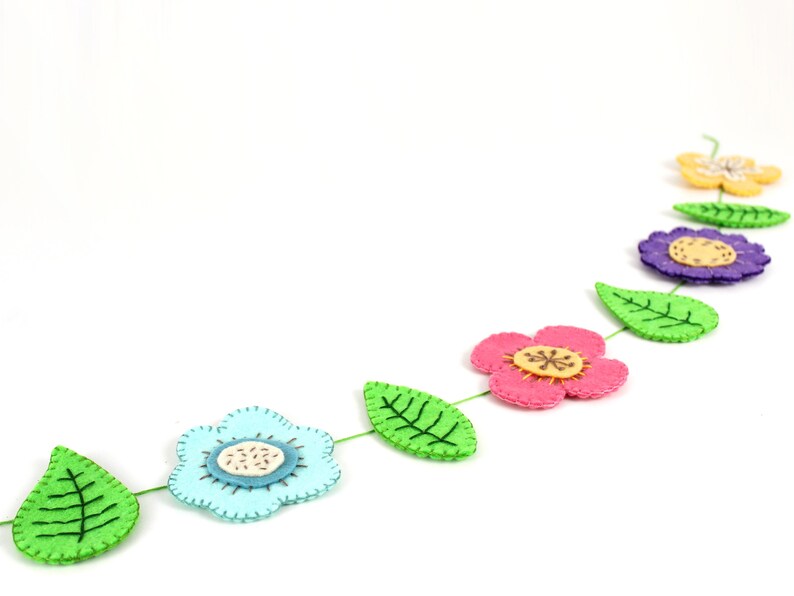 May include: A felt flower garland featuring a variety of colorful flowers and green leaves. The flowers are in shades of pink, blue, purple, and yellow, with detailed stitching and wooden center accents. The garland is strung on a green thread.