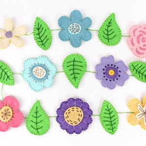 May include: A felt flower garland featuring various colorful flowers and green leaves. The flowers are in shades of pink, blue, purple, and yellow, with detailed stitching. The leaves are a vibrant green. The garland is strung together with green thread.