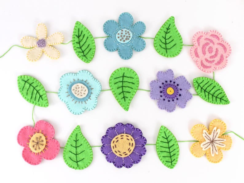 May include: A felt flower garland featuring various colorful flowers and green leaves. The flowers are in shades of pink, blue, purple, and yellow, with detailed stitching. The leaves are a vibrant green. The garland is strung together with green thread.