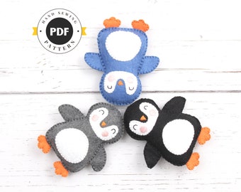 May include: Three felt penguin toys on a white wooden background. The penguins are blue, gray, and black with white bellies and orange feet. The penguins are all lying on their backs with their wings spread out.