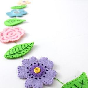 May include: A felt flower garland featuring various colorful flowers and green leaves. The flowers are in shades of pink, blue, and purple, with yellow centers. The leaves are a bright green. The garland is on a white background.