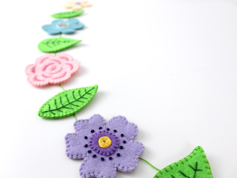 May include: A felt flower garland featuring various colorful flowers and green leaves. The flowers are in shades of pink, blue, and purple, with yellow centers. The leaves are a bright green. The garland is on a white background.