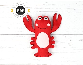 May include: A red felt lobster with black stitching and white felt eyes. The lobster is smiling and has a white felt belly. The lobster is on a white wooden background. The image includes a watermark that says "Hand Sewing PDF Pattern".