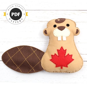 May include: A brown felt beaver with a red felt maple leaf on its chest. The beaver has a brown felt nose and white felt teeth. The beaver's tail is brown felt with a diamond pattern stitched in gold thread. The beaver is on a white wooden background.
