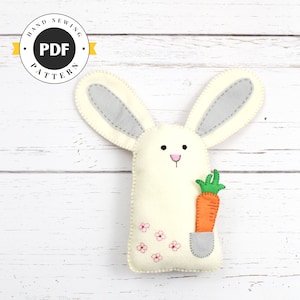 Puede incluir: Un conejito de fieltro blanco con orejas grises que sostiene una zanahoria. El conejito tiene detalles bordados en rosa en la cara y un bolsillo en el costado con flores rosas bordadas. El conejito está sentado sobre un fondo de madera blanco. Patrón PDF para coser a mano.
