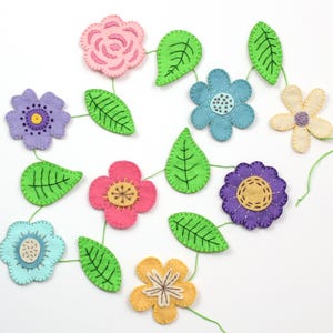 May include: A colorful felt flower garland featuring various flower shapes in pink, purple, blue, and yellow, connected by green leaves and green thread. The flowers have embroidered details and are arranged on a white background.