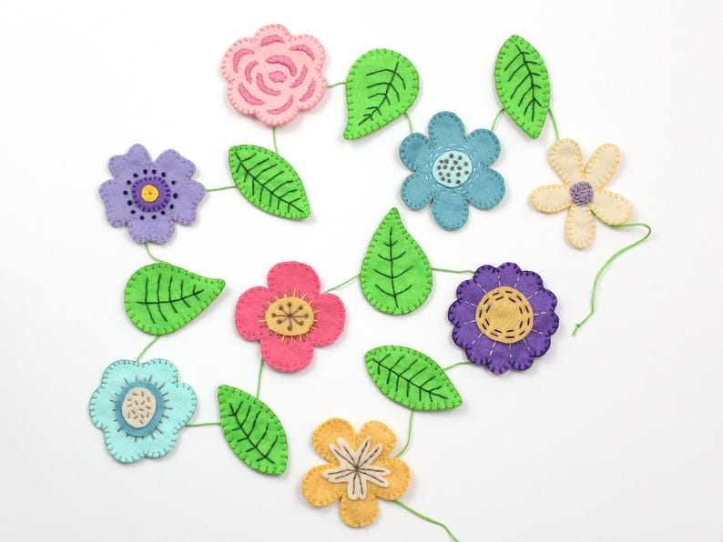 May include: A colorful felt flower garland featuring various flower shapes in pink, purple, blue, and yellow, connected by green leaves and green thread. The flowers have embroidered details and are arranged on a white background.