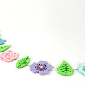 May include: A felt flower garland featuring various colorful flowers and green leaves. The garland includes pink, blue, purple, and yellow flowers, with green leaves. The flowers and leaves are connected by a green string.