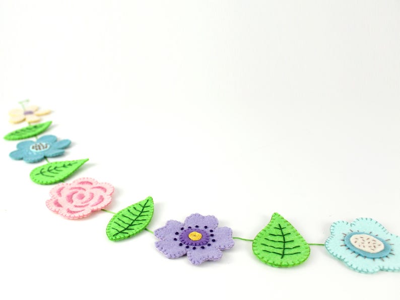 May include: A felt flower garland featuring various colorful flowers and green leaves. The garland includes pink, blue, purple, and yellow flowers, with green leaves. The flowers and leaves are connected by a green string.