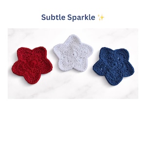 May include: Three crocheted star-shaped coasters in red, white, and blue. The coasters are arranged on a white marble surface. The text "Subtle Sparkle" is above the coasters.