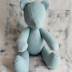 May include: A light blue teddy bear made of textured fabric. The bear has a simple design with stitched features, including small ears and a rounded body. The bear is sitting upright, with its arms and legs extended.