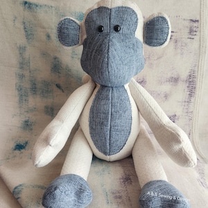 May include: A handmade stuffed monkey in shades of blue and cream. The monkey has a blue face, ears, and feet, with a cream-colored body and limbs. Black button eyes complete the design. The monkey is sitting on a textured surface.