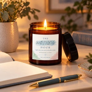 May include: A lit candle in a brown glass jar with a white label featuring the text THE WRITING HOUR, Light when you need to find the words, BERGAMOT & OUD, and THIS LADY WRITES, accompanied by a black lid and a blue pen on a desk.