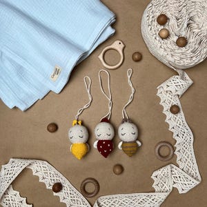 May include: A collection of handmade baby items, including three crocheted dolls with different colored outfits, a light blue blanket, wooden beads, a wooden bird-shaped teether, and cream-colored lace trim.