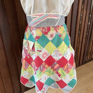 May include: A colorful patchwork apron with a scalloped hem. The apron features a variety of diamond-shaped fabric pieces in red, green, yellow, and floral patterns. The waistband is gray with a geometric design. The apron is displayed on a mannequin.