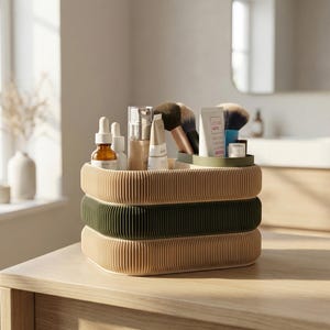 May include: A three-tiered, corrugated paper organizer in beige and olive green, holding beauty products. The top tray contains makeup brushes, tubes, and bottles. The organizer sits on a wooden surface, suggesting bathroom or vanity use.