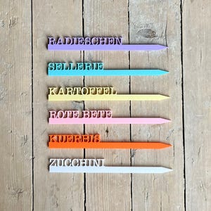 Puede incluir: Marcadores de jardín coloridos con texto en alemán. Los marcadores son de varios colores, incluyendo morado, turquesa, amarillo, rosa, naranja y blanco. Cada marcador tiene una palabra alemana diferente, como "Radieschen" y "Zucchini".