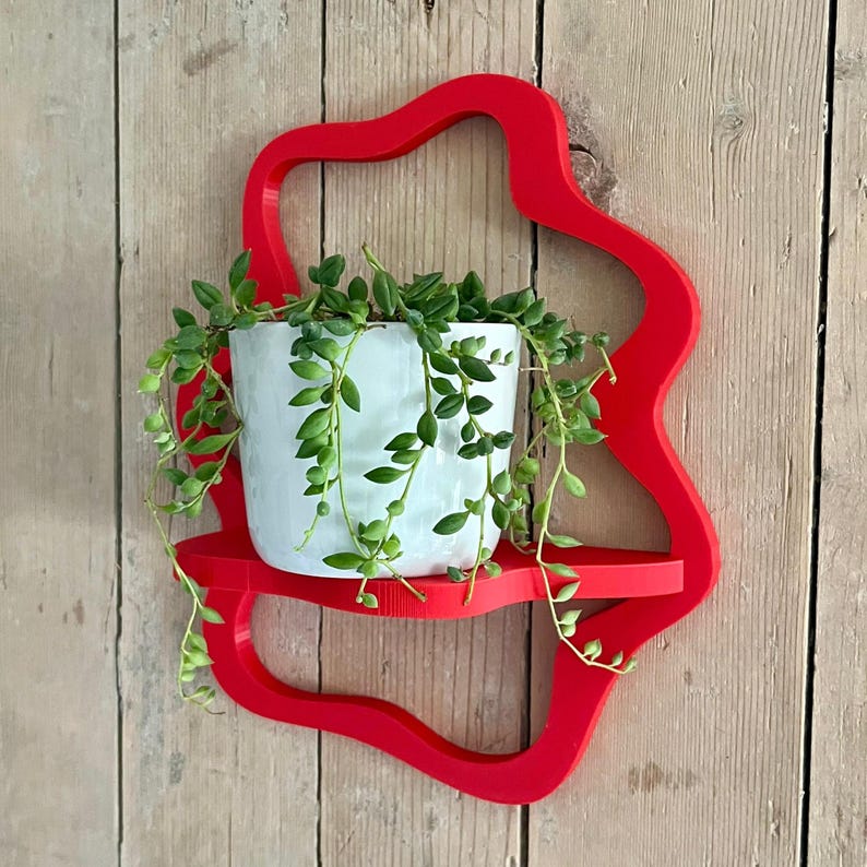 May include: A vibrant red, wall-mounted shelf with a unique, flowing design. A white ceramic pot filled with trailing green plants sits on the shelf. The shelf is mounted on a wooden plank wall, creating a striking contrast.