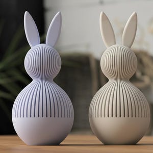 Könnte beinhalten: Zwei dekorative Hasenfiguren. Einer ist hellviolett, der andere beige. Jeder hat einen abgerundeten Körper mit vertikalen Rillen und langen Ohren. Die untere Hälfte jedes Hasen ist einfarbig. Sie stehen auf einer Holzoberfläche.