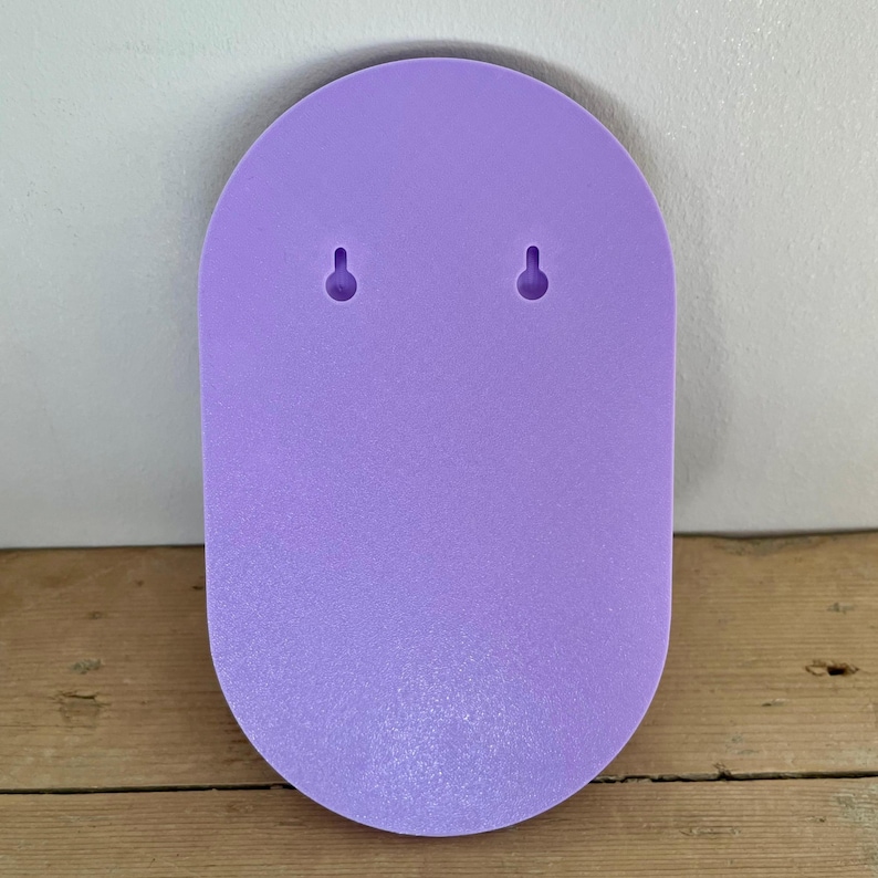 May include: A light purple, oval-shaped wall hanging with two small holes for mounting. The wall hanging is made of a solid, smooth material and has a slightly textured surface. The background is a light gray wall and a wooden surface.