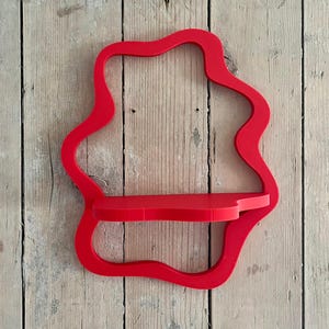May include: A vibrant red wall shelf with a unique, flowing, organic shape. The shelf has a small, rectangular platform for displaying items. The shelf is mounted on a weathered, light-colored wooden wall, adding a touch of rustic charm.