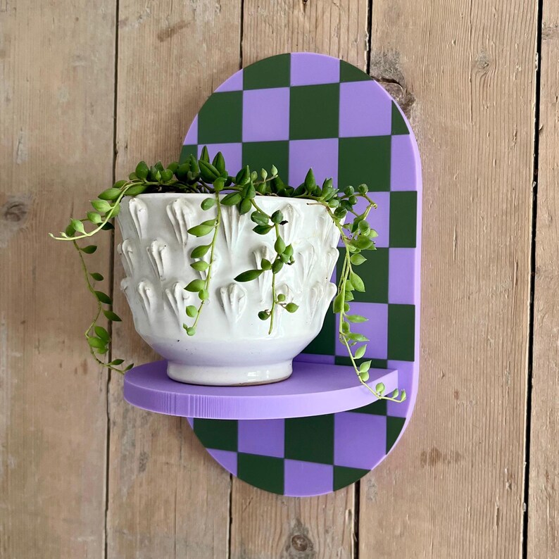 May include: A white ceramic planter with a textured design, filled with trailing green plants, sits on a purple shelf. The shelf is attached to a decorative wall-mounted piece with a checkerboard pattern in green and purple.