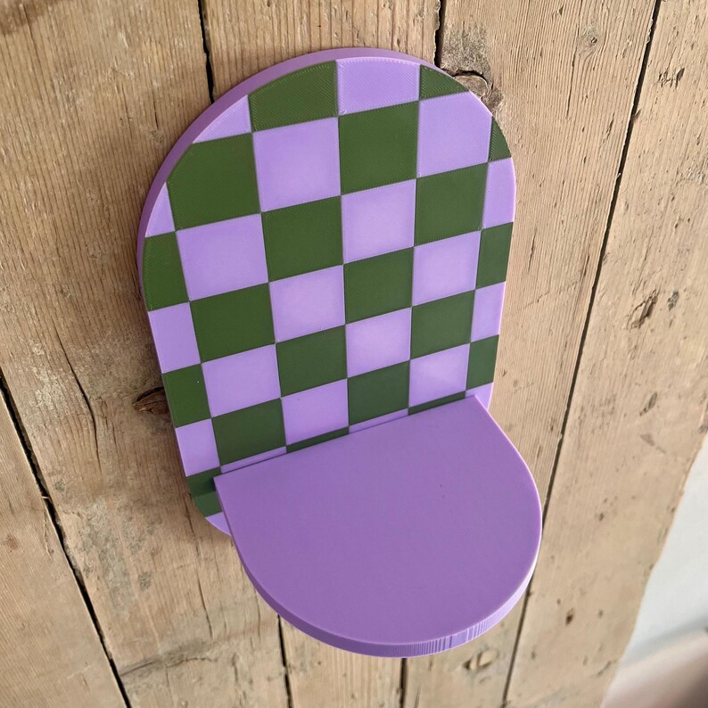 May include: A decorative wall shelf with a checkerboard pattern in green and lavender. The shelf has a semi-circular base and is mounted on a wooden surface. The shelf is approximately 8 inches wide and 10 inches tall.