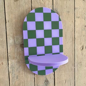May include: A decorative wall shelf with a checkerboard pattern in green and lavender. The shelf has a rounded top and a small, curved shelf extending from the bottom. The shelf is made of a solid material and is mounted on a wooden surface.