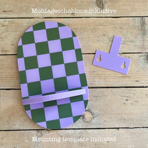 May include: A purple and green checkered phone stand with a matching mounting template. The stand has a shelf and is labeled "Mounting template included". The stand is made of acrylic.