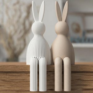 Op de afbeelding: Twee decoratieve konijnenbeeldjes. Eén is wit met verticale lijnen, de andere is beige. Beide hebben lange oren en cilindrische poten, zittend op een houten oppervlak. De achtergrond is wazig, wat wijst op een interieur.