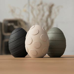 Può includere: Tre uova di Pasqua decorative. L'uovo a sinistra è nero con strisce diagonali. L'uovo centrale è beige con motivi circolari. L'uovo a destra è verde oliva con strisce orizzontali.