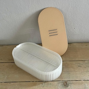 Pode incluir: Um recipiente oval transparente e canelado com uma tampa a condizer. A tampa é de cor bege claro com três ranhuras horizontais. O recipiente está sobre uma superfície de madeira.